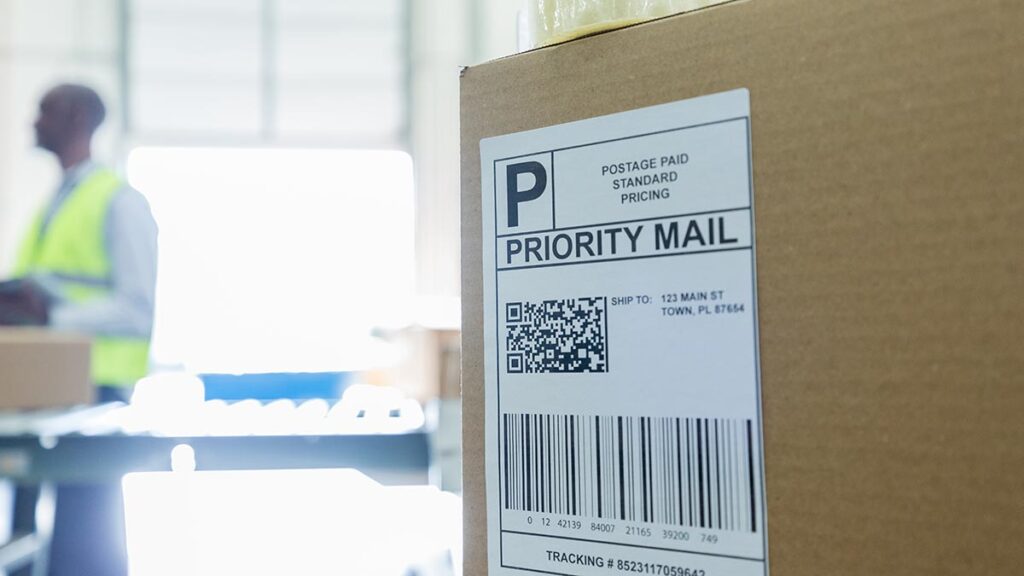 Focus is on a cardboard box in the foreground with a priority mail label.  It is waiting to leave the a warehouse that can be seen in the background.  An employee stands in the background near an open garage door.